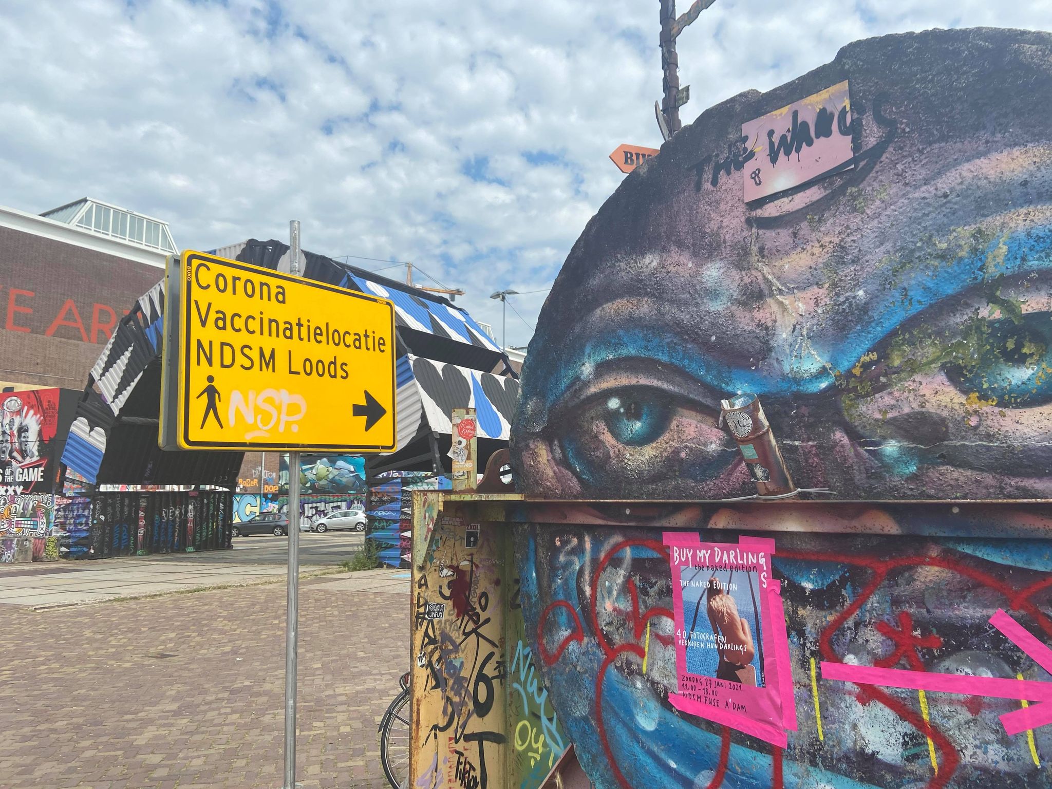 A street sign that states 'Corona Vaccinatielocatie NDSM Loods.' 
            The sign is surrounded by colorful street art and graffiti.